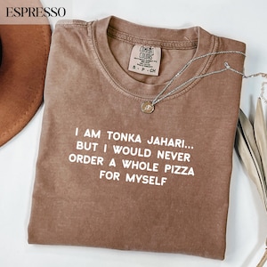May include: Espresso-colored t-shirt with the text "I AM TONKA JAHARI... BUT I WOULD NEVER ORDER A WHOLE PIZZA FOR MYSELF" in white. The shirt has a crew neck and short sleeves. A silver necklace with a pendant is draped across the shirt.