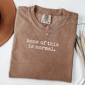 May include: A brown t-shirt with the slogan "none of this is normal" printed in white. A silver necklace with a circular pendant lies on the shirt. The shirt is folded on a white surface.