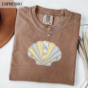 Puede incluir: Camiseta color espresso con un diseño de concha. La concha está hecha de tela estampada azul y amarilla. Un collar plateado con un pequeño colgante está drapeado sobre la camisa. La camisa tiene una etiqueta Comfort Colors.