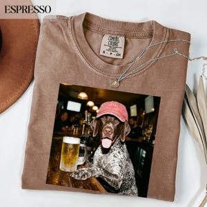 May include: A brown short-sleeved t-shirt with a Comfort Colors label at the neck and a graphic print featuring a brown and white spotted dog wearing a red baseball cap, sitting at a bar next to a glass mug filled with a golden beer.