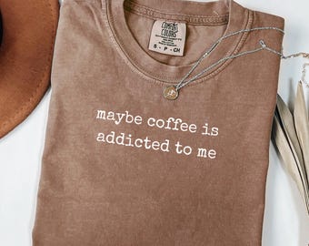 Peut inclure: T-shirt marron avec l'inscription blanche "maybe coffee is addicted to me". Un collier argenté avec un petit pendentif est posé sur le t-shirt. Le t-shirt a un col rond et une étiquette "Comfort Colors".