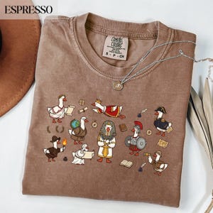Könnte beinhalten: Espressofarbenes Comfort Colors T-Shirt mit einer skurrilen Illustration von Gänsen in historischen Kostümen. Die Gänse werden als römische Senatoren, Pharaonen und Soldaten dargestellt. Eine silberne Halskette mit Anhänger liegt über dem Shirt.