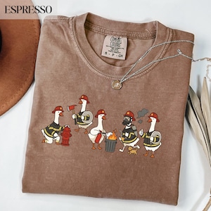 Puede incluir: Una camiseta de manga corta marrón con un gráfico de cinco gansos de dibujos animados vestidos de bomberos. Los gansos llevan cascos rojos, chaquetas con rayas amarillas, una boca de incendio, una manguera, un extintor y un perro pequeño.