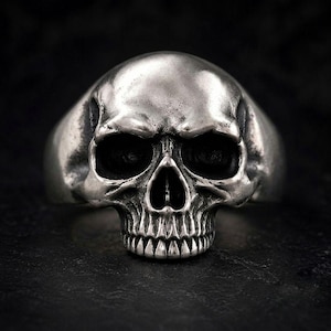 Puede incluir: Un anillo de tono plateado que presenta un diseño detallado de un cráneo humano en tres dimensiones. El metal tiene un acabado pulido en las superficies elevadas y áreas oscuras y hundidas para enfatizar los rasgos anatómicos de las cuencas oculares, la nariz y los dientes.