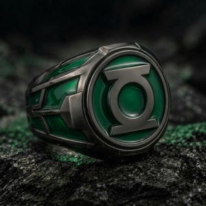 May include: A dark metallic ring featuring a circular face with the Green Lantern symbol in relief. The design includes a vibrant green enamel inlay surrounding the central emblem and extending along the sides of the band with geometric cutouts.