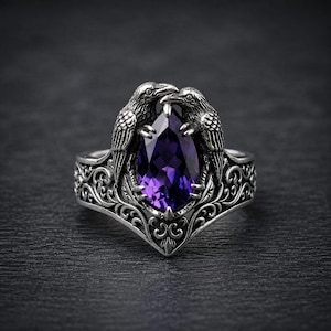 May include: A silver ring featuring a pear-shaped, deep purple gemstone held by two detailed raven figures. The band is crafted with intricate, swirling metal filigree patterns that extend from the base up to the central stone setting.