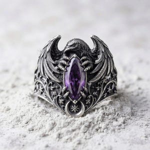 May include: A silver-toned ring featuring a detailed raven with spread wings clutching a marquise-cut purple gemstone in its talons. The band is decorated with intricate floral scrollwork and a small crescent moon symbol with a star at the base.