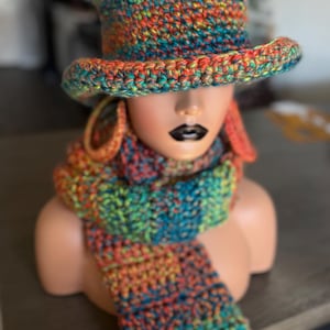 Puede incluir: Un conjunto de sombrero de copa y bufanda a juego tejidos a ganchillo con un patrón de hilo jaspeado en tonos vibrantes de naranja, azul, verde y amarillo. El sombrero tiene una copa estructurada y un ala ancha ligeramente curvada hacia arriba.