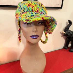 Puede incluir: Un vibrante gorro de ganchillo multicolor con visera y pendientes de aro a juego. El gorro y los pendientes presentan una mezcla de hilo amarillo ne&oacute;n, azul, morado y rosa. El gorro se exhibe en una cabeza de maniqu&iacute;.