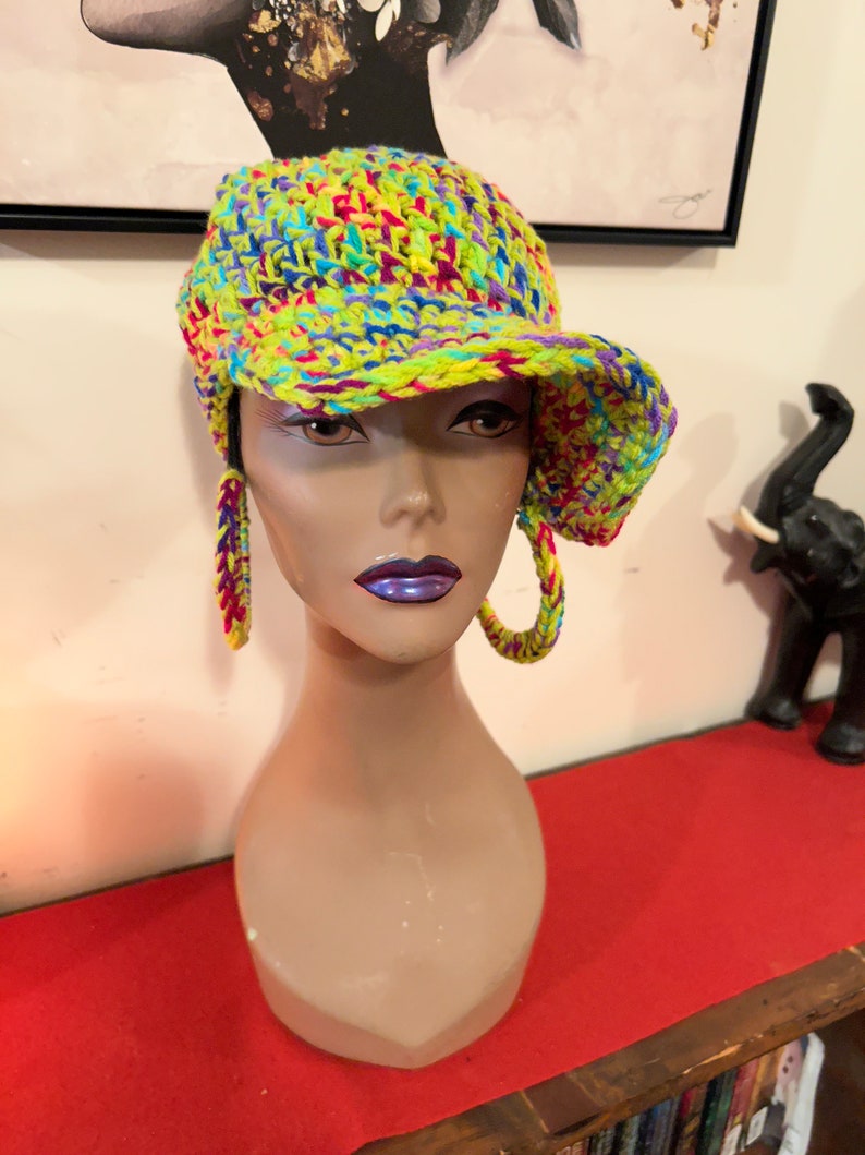 Puede incluir: Un vibrante gorro de ganchillo multicolor con visera y pendientes de aro a juego. El gorro y los pendientes presentan una mezcla de hilo amarillo ne&oacute;n, azul, morado y rosa. El gorro se exhibe en una cabeza de maniqu&iacute;.