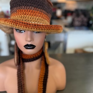 Puede incluir: Un conjunto de sombrero y bufanda de ganchillo con rayas horizontales en tonos marrón oscuro, marrón medio y naranja dorado. El sombrero tiene un ala ancha y una copa alta, mientras que la bufanda larga está hecha del mismo hilo texturizado.