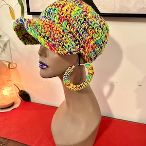 Puede incluir: Un sombrero vibrante tejido a mano con visera y pendientes de aro a juego. El sombrero y los pendientes presentan una colorida mezcla de hilo verde ne&oacute;n, rosa, azul y amarillo. Los accesorios se exhiben en una cabeza de maniqu&iacute;.