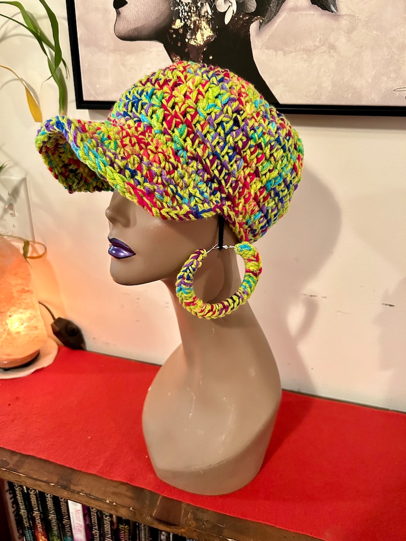 Puede incluir: Un sombrero vibrante tejido a mano con visera y pendientes de aro a juego. El sombrero y los pendientes presentan una colorida mezcla de hilo verde ne&oacute;n, rosa, azul y amarillo. Los accesorios se exhiben en una cabeza de maniqu&iacute;.