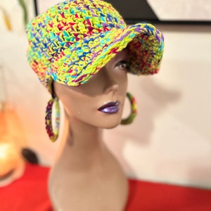 Puede incluir: Una gorra de b&eacute;isbol de ganchillo de colores brillantes en tonos amarillo ne&oacute;n, azul, morado y rosa. La gorra se exhibe en una cabeza de maniqu&iacute;, mostrando su dise&ntilde;o texturizado y su vibrante paleta de colores. La visera del sombrero est&aacute; ligeramente curvada.