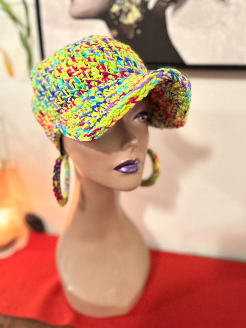 Puede incluir: Una gorra de b&eacute;isbol de ganchillo de colores brillantes en tonos amarillo ne&oacute;n, azul, morado y rosa. La gorra se exhibe en una cabeza de maniqu&iacute;, mostrando su dise&ntilde;o texturizado y su vibrante paleta de colores. La visera del sombrero est&aacute; ligeramente curvada.
