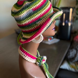 May include: A colorful crochet hat and matching necklace set. The hat features horizontal stripes in shades of green, pink, cream, and blue. The necklace has a flower accent and a long, striped tie. The set is displayed on a mannequin head.