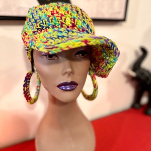 Puede incluir: Un sombrero de ganchillo colorido con visera, con una vibrante mezcla de hilo amarillo, azul, rojo y morado. El sombrero se muestra en una cabeza de maniqu&iacute;, combinado con pendientes de aro a juego. El estilo general es hecho a mano y &uacute;nico.