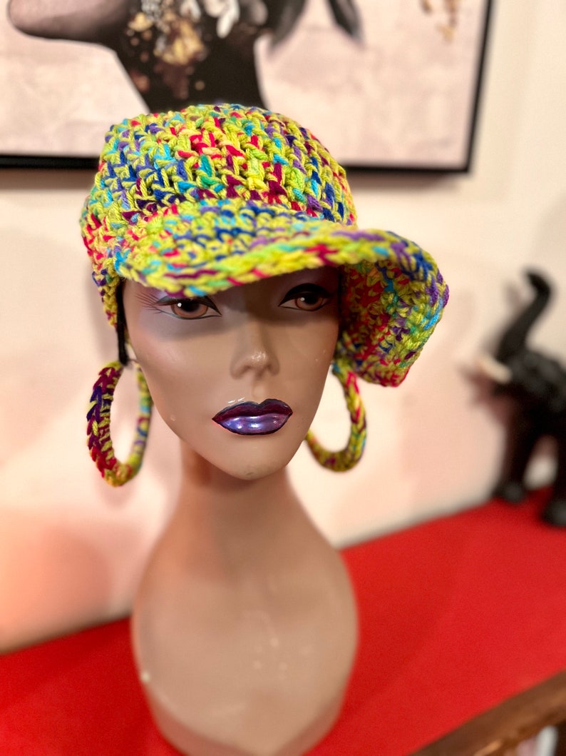 Puede incluir: Un sombrero de ganchillo colorido con visera, con una vibrante mezcla de hilo amarillo, azul, rojo y morado. El sombrero se muestra en una cabeza de maniqu&iacute;, combinado con pendientes de aro a juego. El estilo general es hecho a mano y &uacute;nico.