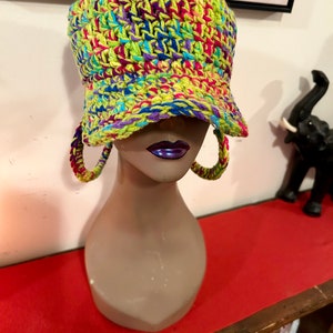 Puede incluir: Un sombrero de pescador tejido a mano con colores vibrantes, hecho con hilo verde ne&oacute;n, rosa, azul y morado. El sombrero tiene un ala peque&ntilde;a y se exhibe en una cabeza de maniqu&iacute; con pendientes de aro tejidos a juego. Un accesorio de moda &uacute;nico.