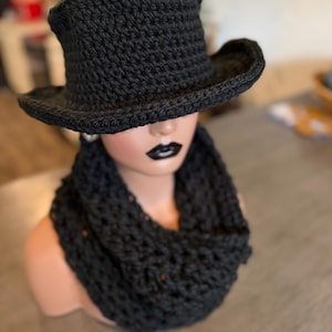 Puede incluir: Un sombrero de copa negro tejido a ganchillo con un ala ancha ligeramente levantada y una bufanda infinita a juego también tejida a ganchillo. El sombrero tiene una copa cilíndrica estructurada y la bufanda un patrón de punto grueso.