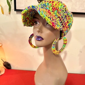 Puede incluir: Un busto de maniqu&iacute; exhibe un sombrero tejido a mano con colores vibrantes. El sombrero presenta una mezcla de hilo amarillo ne&oacute;n, rosa, azul y morado. Los pendientes de aro a juego, tambi&eacute;n tejidos con los mismos colores brillantes, completan el look.