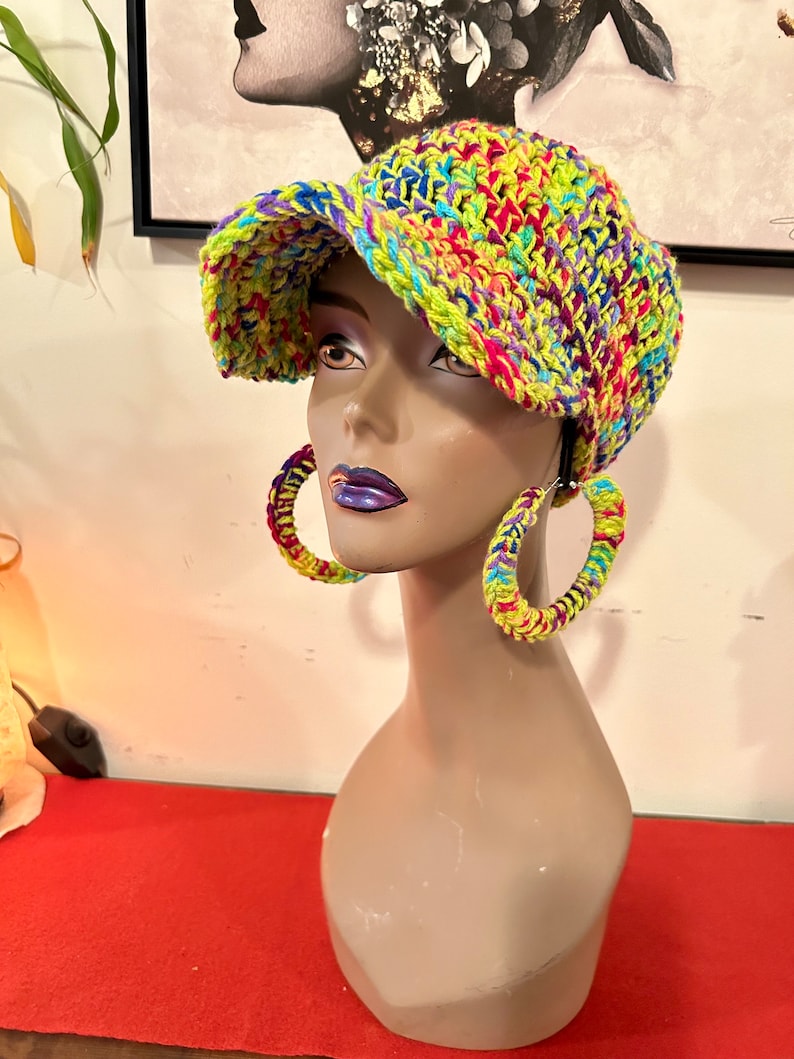 Puede incluir: Un busto de maniqu&iacute; exhibe un sombrero tejido a mano con colores vibrantes. El sombrero presenta una mezcla de hilo amarillo ne&oacute;n, rosa, azul y morado. Los pendientes de aro a juego, tambi&eacute;n tejidos con los mismos colores brillantes, completan el look.