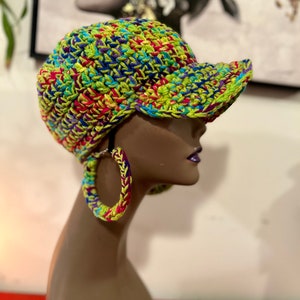 Puede incluir: Gorra de b&eacute;isbol de ganchillo colorida con una visera curva, que presenta una vibrante mezcla de hilos verdes, azules, amarillos y rosas. La gorra se muestra en una cabeza de maniqu&iacute;, combinada con pendientes de aro de ganchillo a juego. El estilo es hecho a mano y &uacute;nico.
