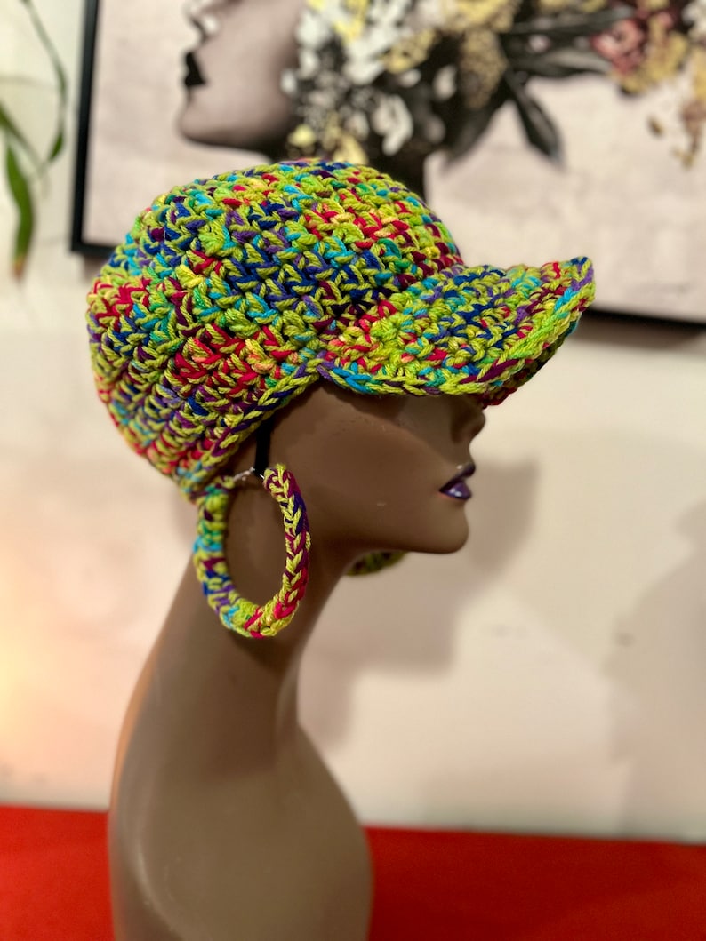 Puede incluir: Gorra de b&eacute;isbol de ganchillo colorida con una visera curva, que presenta una vibrante mezcla de hilos verdes, azules, amarillos y rosas. La gorra se muestra en una cabeza de maniqu&iacute;, combinada con pendientes de aro de ganchillo a juego. El estilo es hecho a mano y &uacute;nico.