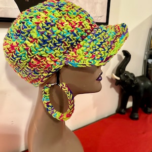Puede incluir: Una gorra de b&eacute;isbol vibrante, tejida a mano, en una mezcla de hilo verde ne&oacute;n, azul, morado, rosa y amarillo. Tambi&eacute;n se ven pendientes de aro de ganchillo a juego. La gorra tiene una visera estructurada y un dise&ntilde;o colorido.