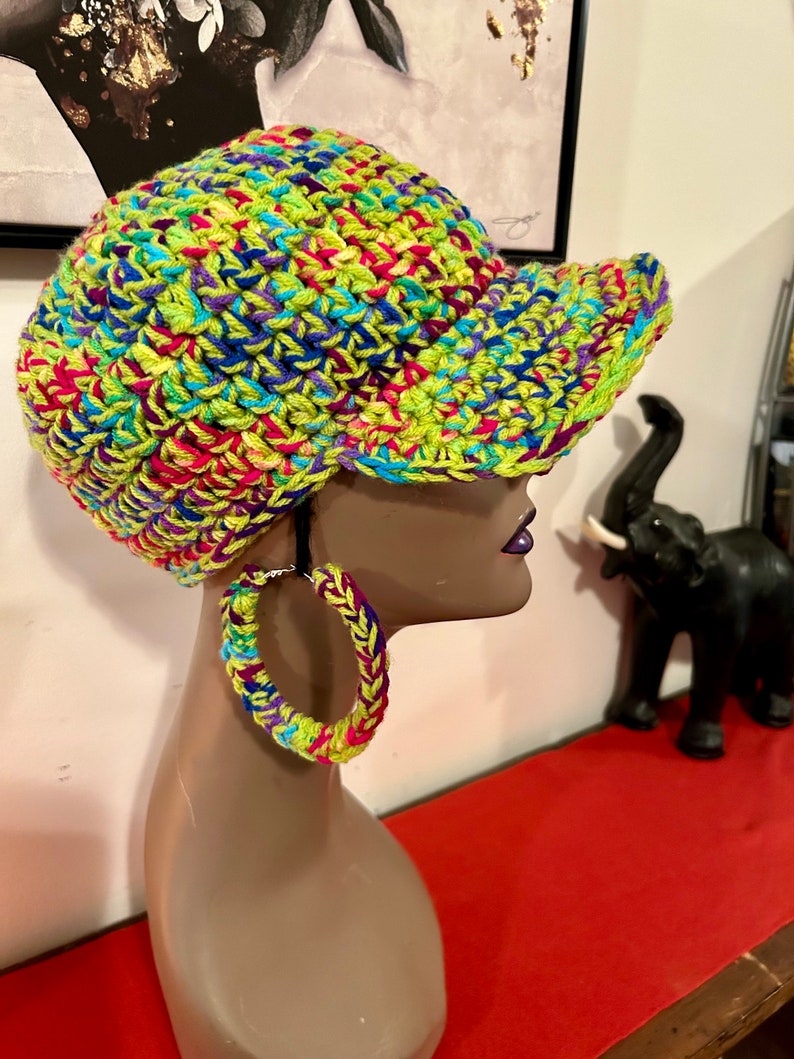 Puede incluir: Una gorra de b&eacute;isbol vibrante, tejida a mano, en una mezcla de hilo verde ne&oacute;n, azul, morado, rosa y amarillo. Tambi&eacute;n se ven pendientes de aro de ganchillo a juego. La gorra tiene una visera estructurada y un dise&ntilde;o colorido.