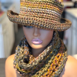 Puede incluir: Un conjunto de sombrero y bufanda infinita de ganchillo con un patrón de hilo jaspeado en tonos verde oliva, amarillo mostaza, marrón y crema. El sombrero tiene una copa estructurada y ala ancha, mientras que la bufanda es de punto grueso.