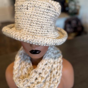 Puede incluir: Un sombrero de copa de ganchillo color crema con sutiles motas oscuras y una bufanda infinita de punto grueso a juego. El sombrero presenta una copa plana y un ala ancha ligeramente levantada, mientras que la bufanda tiene un tejido abierto.