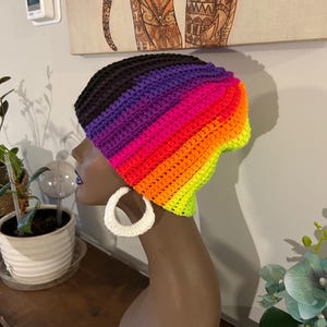 Puede incluir: Un gorro de punto de crochet colorido con rayas de color negro, morado, rosa, naranja, amarillo y verde. El gorro está en una cabeza de maniquí.