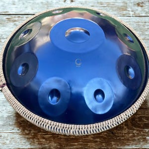 May include: A blue steel handpan instrument with a circular design and multiple sound holes. The instrument has a protective woven cover around the edge. The word "Foset" is printed on the surface.