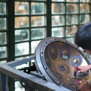 May include: A person working on a steel pan drum. The instrument is bronze-colored with multiple circular indentations. The drum is mounted on a gray metal stand, with a window in the background.
