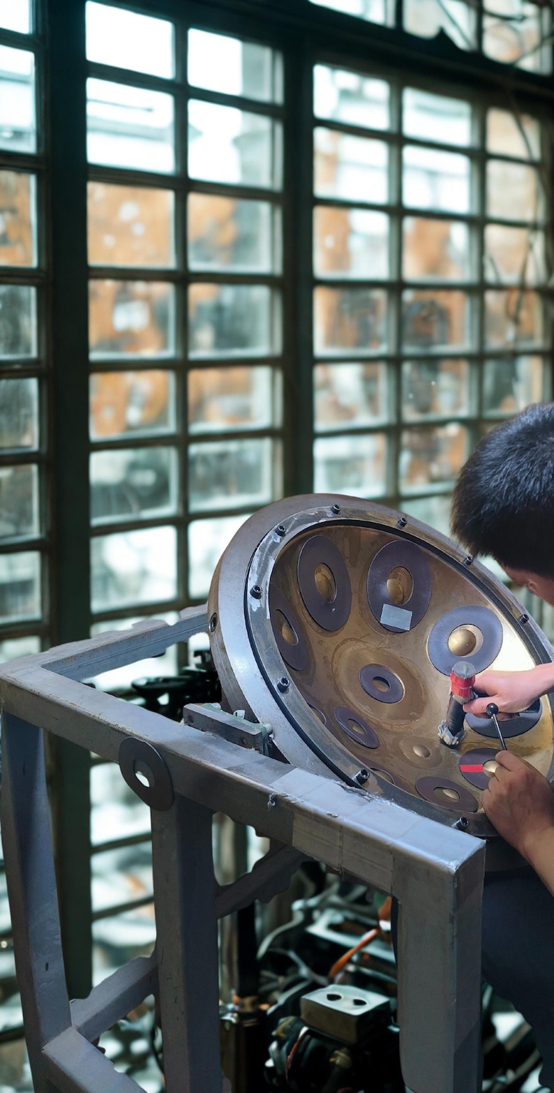 May include: A person working on a steel pan drum. The instrument is bronze-colored with multiple circular indentations. The drum is mounted on a gray metal stand, with a window in the background.