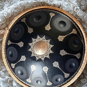 May include: A black steel handpan drum featuring a central gold-toned dome and intricate gold filigree patterns radiating outward. The instrument has multiple circular tone fields and a textured gold-colored rim encircling the entire edge.