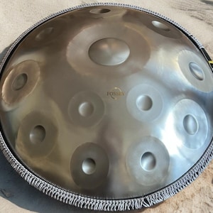 May include: A silver-colored handpan instrument with multiple circular indentations across its surface. The word "FOSSEY" is printed on the instrument. The handpan is surrounded by a gray and black fringed border.