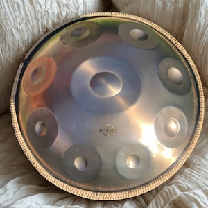 Könnte beinhalten: Ein silbernes Handpan-Instrument mit goldenem Rand und dem Wort "FOSSEY" auf der Oberfläche. Das Handpan hat mehrere kreisförmige Vertiefungen und liegt auf einer weißen, strukturierten Oberfläche.
