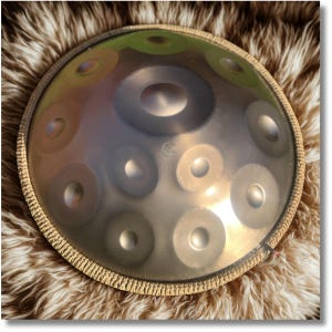 May include: A steel handpan instrument with a circular shape and multiple circular sound holes. The instrument has a bronze-like finish and is surrounded by a woven fabric edge. The background is a fluffy, brown and white fur.