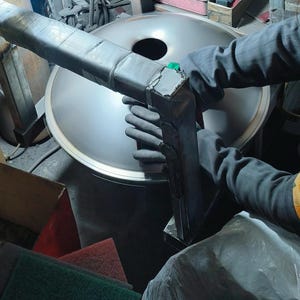 May include: A person wearing black gloves is working on a large, silver metal object with a dark, rectangular tool. The tool is attached to the metal object. The background includes other tools and materials.
