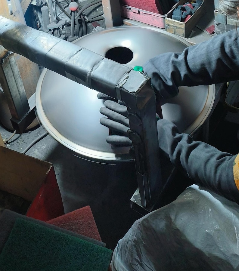 May include: A person wearing black gloves is working on a large, silver metal object with a dark, rectangular tool. The tool is attached to the metal object. The background includes other tools and materials.
