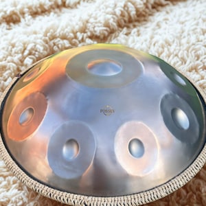 May include: A silver handpan instrument with multiple circular sound holes. The handpan has a metallic sheen with a woven border. The word "FOSSEY" is printed on the surface. The instrument is designed for musical performance.
