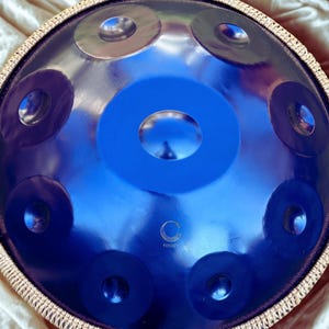 May include: A blue handpan instrument with a shiny, reflective surface. The handpan features a central blue circle surrounded by smaller, dark blue circles. Silver-colored metal rings are also visible. The instrument has a circular shape and is framed by a decorative border.