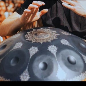May include: A dark blue handpan drum with a decorative gold and white floral pattern. The instrument has several circular sound holes and is being played by a person wearing a black shirt. The rim is a woven brown material.