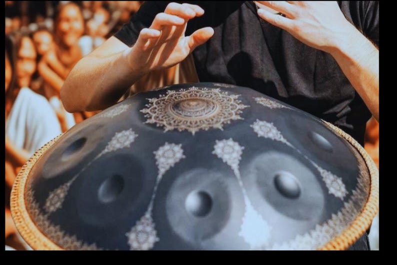 May include: A dark blue handpan drum with a decorative gold and white floral pattern. The instrument has several circular sound holes and is being played by a person wearing a black shirt. The rim is a woven brown material.