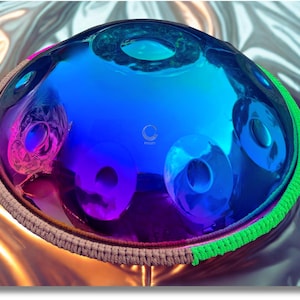 May include: A vibrant, iridescent blue handpan instrument with a polished, reflective surface. The handpan features several circular sound holes and is trimmed with a green and brown woven border. The instrument reflects pink and purple hues.