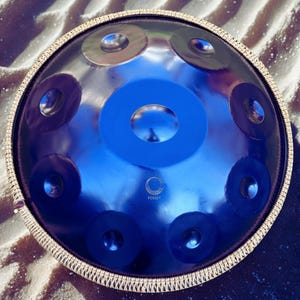May include: A blue handpan instrument with a shiny, reflective surface. The handpan has a central blue circle and several smaller, dark blue and silver-colored circular indentations. The rim is trimmed with a decorative, woven material. The brand name "FOSSEY" is visible.