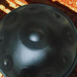 May include: A dark gray steel handpan instrument with multiple circular sound holes. The handpan has a central indentation and a logo near the bottom. The instrument is set against a patterned, reddish-brown background.