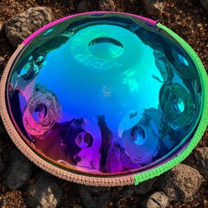 May include: A vibrant, iridescent steel handpan drum with a rainbow color scheme. The instrument has a central note and several surrounding tone fields. The rim is protected by a woven, multi-colored band.